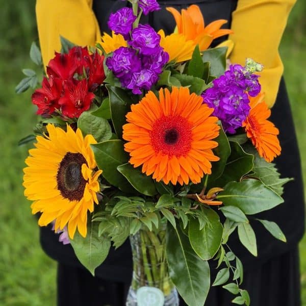 Vibrant mixed flower bouquet featuring sunflowers, gerbera daisies, and lisianthus in a glass vase.