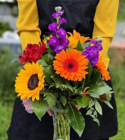 1. posy in a vase Vibrant mixed flower bouquet with sunflowers, gerbera daisies, and purple blooms in a glass vase.