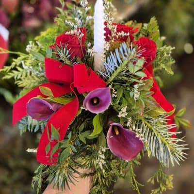Vibrant floral holiday bouquet with red roses, purple calla lilies, greenery, and a white candle.