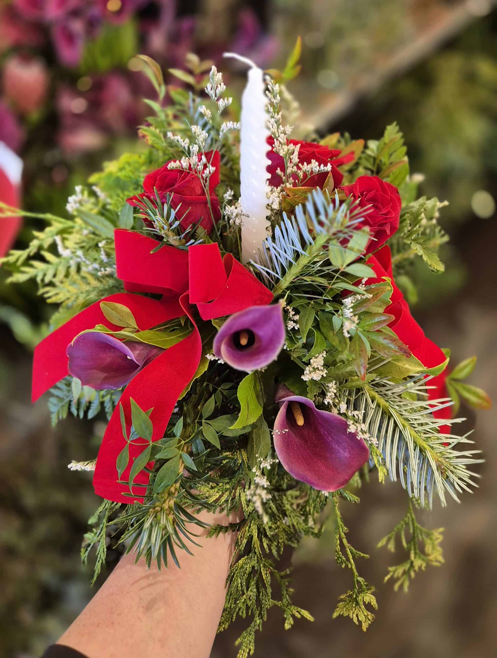 Vibrant floral holiday bouquet with red roses, purple calla lilies, greenery, and a white candle.