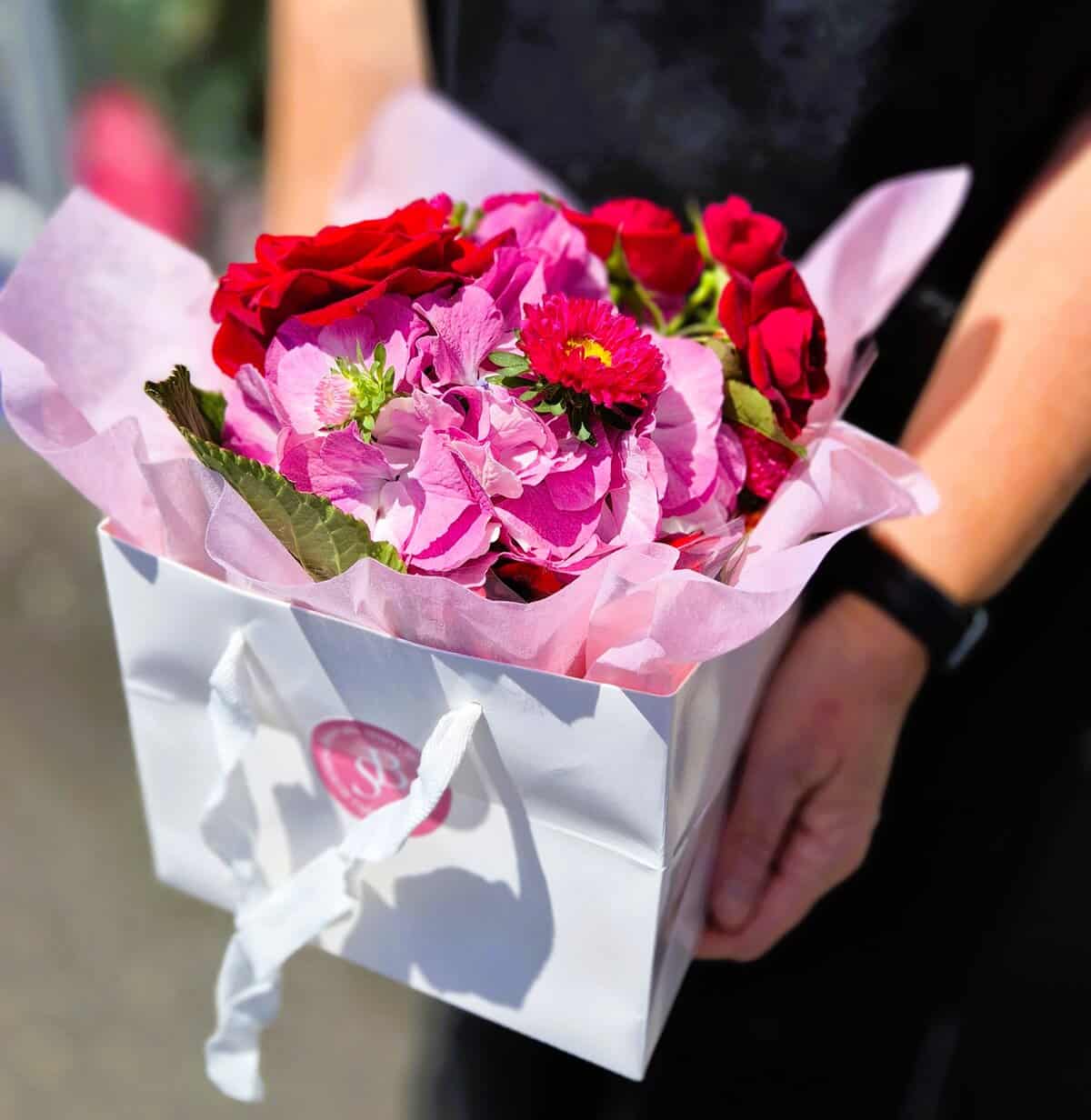 A vibrant flower bouquet featuring pink and red blossoms, beautifully arranged in a white gift bag with tissue paper, perfect for any special occasion.