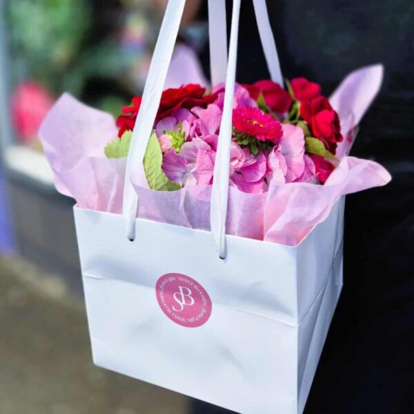 Vibrant pink floral arrangement in white gift bag with Simply Blooming branding, fresh flowers for special events.