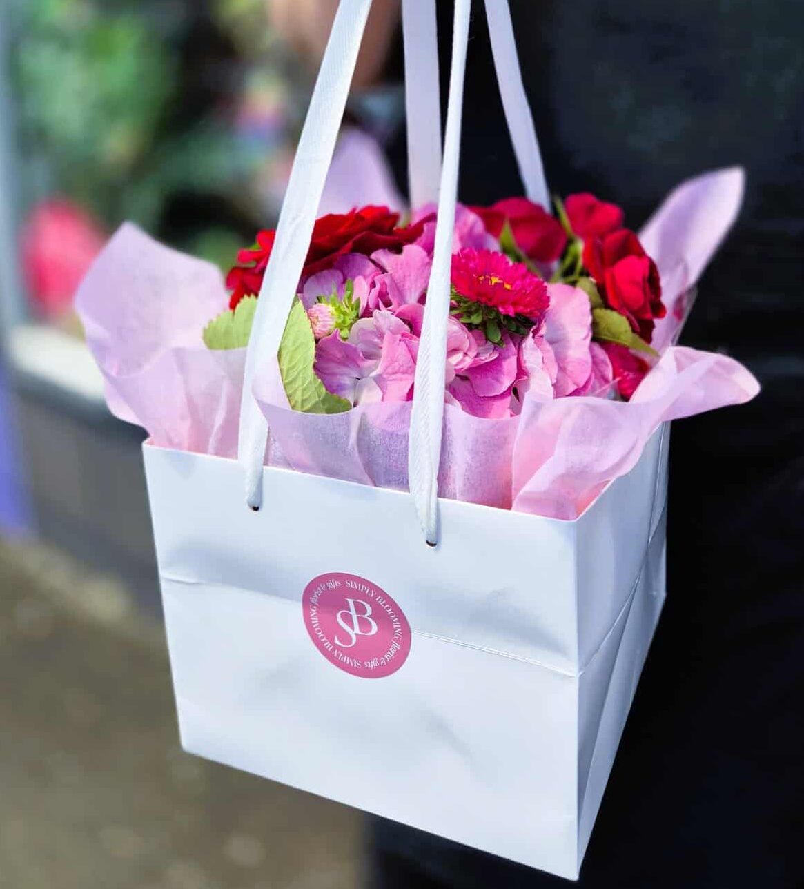 Vibrant pink floral arrangement in white gift bag with Simply Blooming branding, fresh flowers for special events.