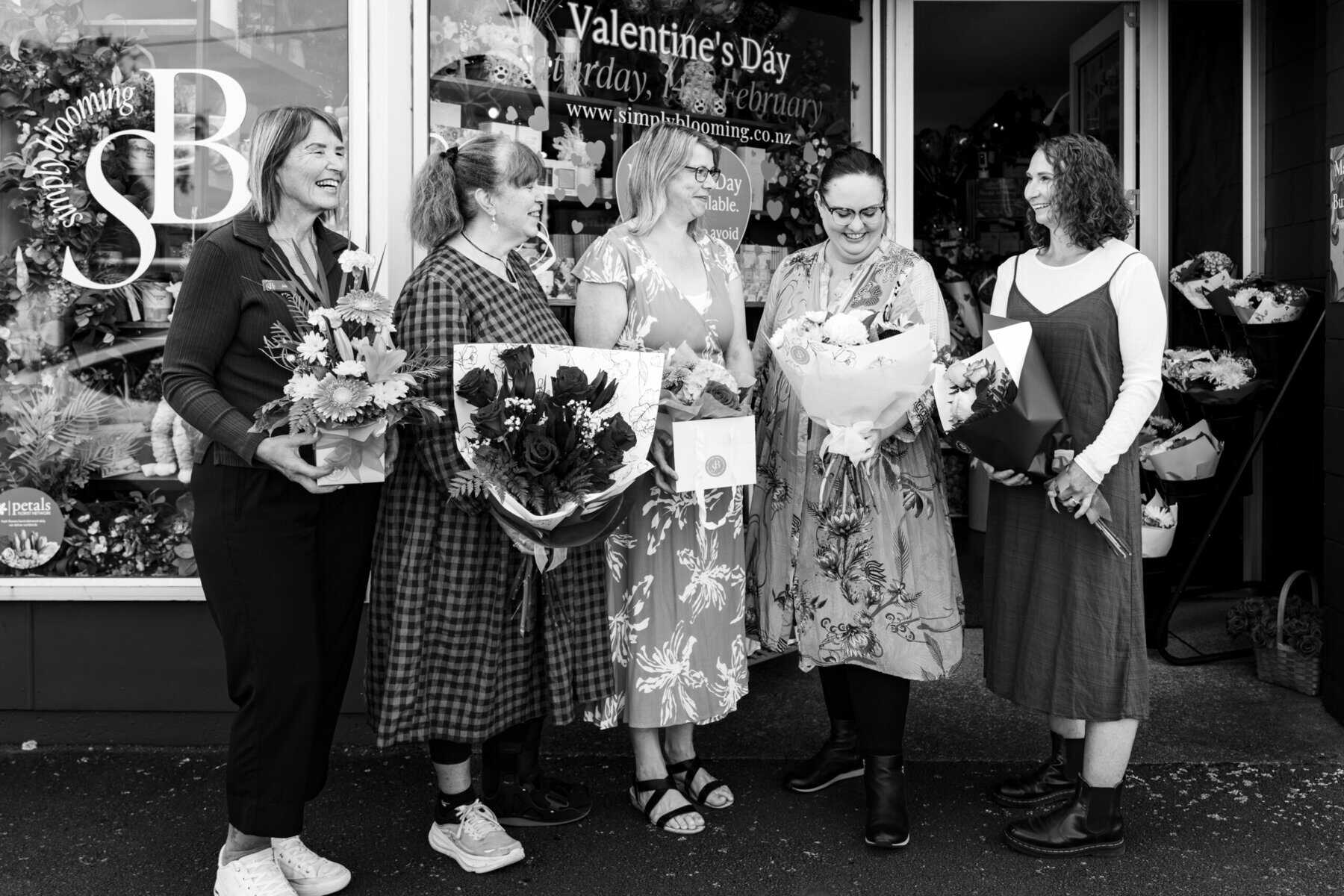 Group of women holding bouquets outside Simply Blooming store, celebrating teamwork.