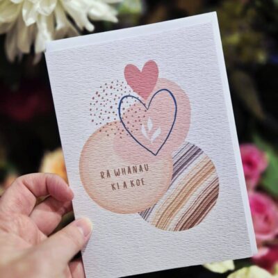 Hand holding a greeting card with heart illustrations and Maori message, floral background.
