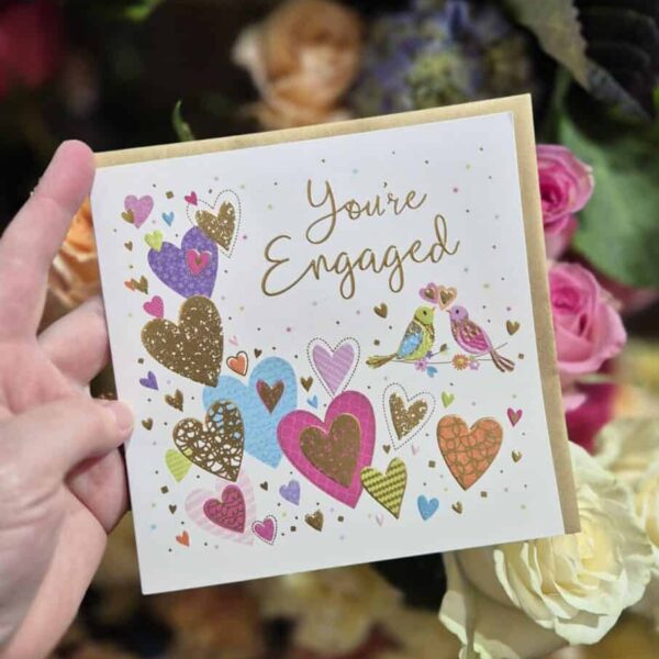 A hand holds a vibrant engagement card decorated with hearts and birds, surrounded by flowers.